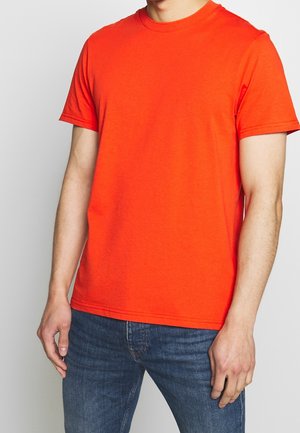Homme portant un t-shirt orange vif uni à manches courtes et un jean en denim bleu, debout devant un fond neutre.