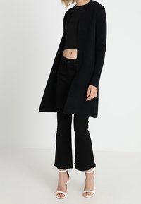 Zwarte lange cardigan, gemêleerde stof, open voorkant, gedragen over een zwarte cropped top en hoge zwarte flared jeans, gecombineerd met witte hakken met banden.