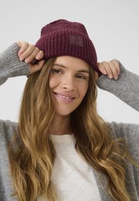 Beanie a coste color borgogna con finitura testurizzata e un'etichetta quadrata in tessuto; indossato con un maglione grigio chiaro e una camicia bianca.