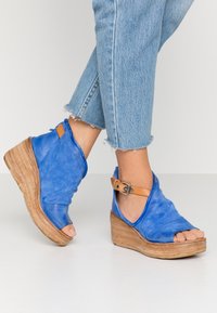 Nohy v modrých semišových sandálech na klínu s tanovými kotníkovými popruhy, doplněné o světle modré džíny se třásněmi na bílém podlaze.
