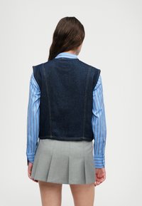 Denimová vesta v tmavomodrej farbe, s surovými okrajmi a viditeľnými švovými detailmi, nosená cez svetlomodrú pruhovanú košeľu a šedú skladanú sukňu.