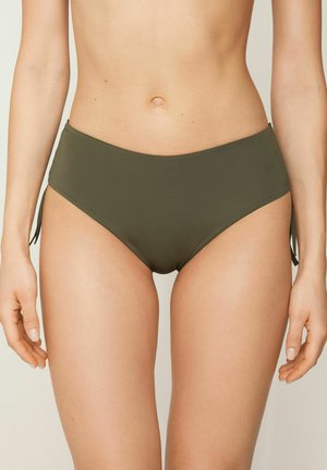 Buste féminin portant un bas de bikini vert olive avec des liens sur les côtés, debout contre un fond beige uni, mains détendues le long du corps.