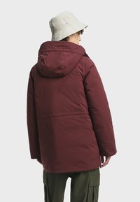 Bordeauxkleurige gewatteerde jas met een capuchon, met een gladde afwerking en een ontspannen pasvorm. Draag het met een lichtgekleurde bucket hat en olijfgroene broek.