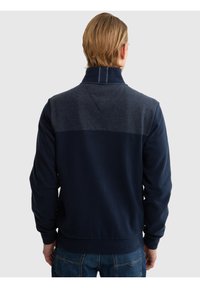 Man met kort blond haar, gekleed in een marineblauw en donkergrijs jack met lange mouwen, gezien van achteren, staand tegen een lichte achtergrond.