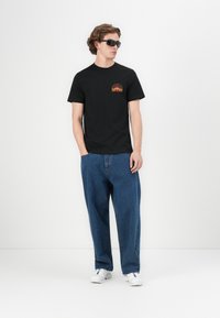 Sort bomulds T-shirt med et farverigt grafisk print på venstre bryst, parret med løse blå denim jeans og hvide sneakers.