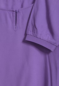 Vêtement violet avec fermeture à boutons, poignets de manches élastiqués et texture de tissu lisse. Design simple avec un éclat subtil.