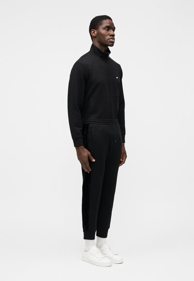 Svart hettejakke med glidelås og høy krage, kombinert med matchende joggebukser med svarte fløyelsdetaljer på sidene. Hvite joggesko fullfører antrekket.