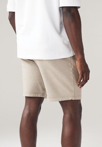 Pantaloni corti in corda beige con taglio dritto, caratterizzati da una sottile texture e dettagli di cucitura, abbinati a una t-shirt bianca di taglio morbido.
