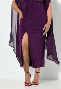 Fialové šaty s volně splývajícím překladem, s nashromážděným živůtkem a vysokým rozparkem na stehno, doplněné o pásek sandálů ve stejném odstínu.