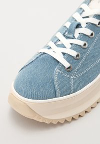 Sneaker in denim blu con lacci bianchi, dotato di occhielli argentati e suola in gomma spessa beige con un motivo a zigzag.