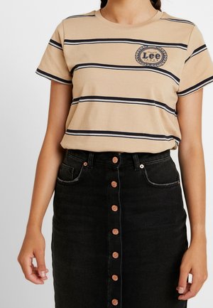 Vrouw draagt een beige gestreept Lee T-shirt, ingestopt in een zwarte spijkerrok met hoge taille en knopen aan de voorkant, staand voor een effen achtergrond.