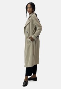 Beige trenchcoat med dubbla knappar, breda slag och sidofickor, med svarta detaljer. Bärs över svarta byxor och loafers.