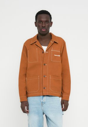 Chaqueta marrón con botones, dos grandes bolsillos frontales, detalles de costura blanca y un pequeño logo en el pecho. Combinada con vaqueros azul claro.