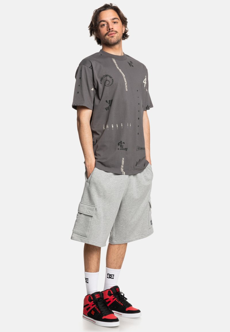 Grijze grafische t-shirt met zwarte print, gecombineerd met lichtgrijze cargo shorts met grote zakken en zwart-rode hoge sneakers.