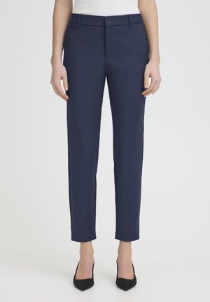 Ženska, ki nosi mornarsko modre hlače s pravimi hlačnicami in črne čeveljce z nabrušenim koncem, stoji ob preprostem svetlem ozadju.