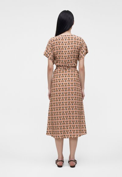 Femme debout, dos tourné au spectateur, portant une robe midi à manches courtes à motifs et des sandales noires sur un fond clair uni.