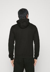 Uomo con una felpa nera con cappuccio e pantaloni neri, in piedi di spalle contro uno sfondo grigio chiaro uniforme.