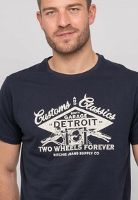 T-shirt en coton bleu marine avec un graphisme de moto vintage et un design textuel en crème. Encolure arrondie, manches courtes et coupe décontractée.