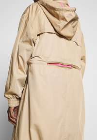 Henkilö, joka on puetutunut beigeen huppariin, jossa on elastiset hihansuut ja puoliksi avattu takatasku, joka paljastaa kirkkaan vaaleanpunaisen vuoren.