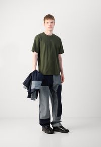 Grünes Baumwoll-T-Shirt mit kurzen Ärmeln. Trägt eine zweifarbige Patchwork-Jeanshose und hält eine dunkelblaue Jeansjacke. Schwarze Sneakers.