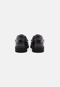 Chaussures en cuir noir avec une texture lisse et une semelle épaisse. Présentent deux sangles décoratives et un petit logo sur le talon.