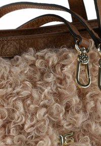 Bolso de mano beige difuso con textura rizada, correas de cuero marrón y acentos en dorado. Presenta un detalle del logo cerca de la parte inferior.