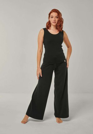 Frau mit roten Haaren, die ein schwarzes ärmelloses Oberteil und weite schwarze Hosen trägt, steht barfuß vor einem schlichten hellen Hintergrund.