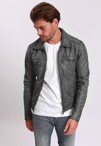 Veste en cuir synthétique grise avec une fermeture éclair à l'avant, deux poches poitrine et un col. Portée par-dessus un t-shirt blanc et associée à un jean en denim clair.
