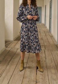 Robe à manches longues, en tissu à volants, avec un motif abstrait bleu marine, crème et beige. Portée avec des bottines olive à la cheville. Sol en planches de bois. Arrière-plan simple.