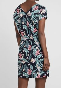 Kobieta w sukience z krótkim rękawem, do kolan, na ciemnym tle z kolorowym tropikalnym wzorem kwiatowym.