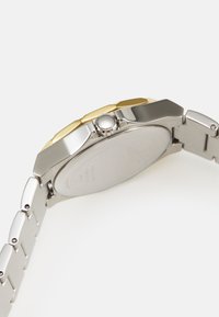 Silver- och guldtonad klocka med ett armband i rostfritt stål. Oktagonal klockkåpa, rund krona och platt, slät baksida med ingraverade detaljer.