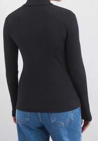 Person som bär en figurnära svart ribbad långärmad topp och blå denimjeans, visad bakifrån mot en enkel ljus vägg.