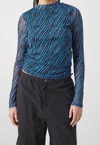 Långärmad topp i genomskinlig blå nät med svarta zebraränder. Har en rundad hals och rynkad detalj vid midjan. Matchas med svarta byxor.