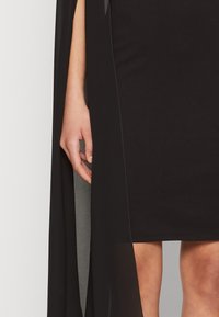 Μαύρο φόρεμα με εφαρμοστή σιλουέτα, που διαθέτει ημιδιαφανές επικάλυμμα τύπου κάπας. Λείο ύφασμα, μήκος έως το γόνατο, και διακριτές ραφές.