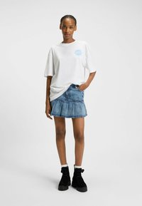 Witte oversized t-shirt met blauwe zonafbeelding; gecombineerd met een denim plissérok en zwarte enkellaarsjes.