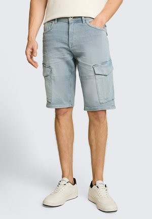 Mann trägt hellblaue Denim-Cargoshorts und weiße Sneakers, steht mit einer Hand in der Tasche vor einem einfarbigen Hintergrund.