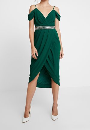 Vrouw draagt een donkergroene cold-shoulder wikkeljurk met een zilveren riem en witte hoge hakken, staand tegen een effen achtergrond.