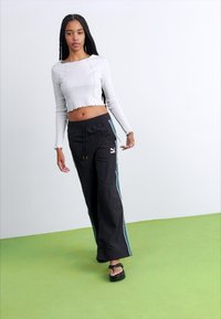 Black drawstring pants with vertical teal stripes, paired with a fitted light gray long-sleeve top. Model stands on green floor, wearing sandals.