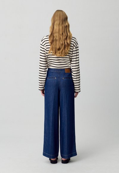 Mujer con cabello rubio largo y ondulado se encuentra de frente a una pared gris claro, vestida con una camiseta de mangas largas de rayas blancas y negras y jeans de pierna ancha azul.