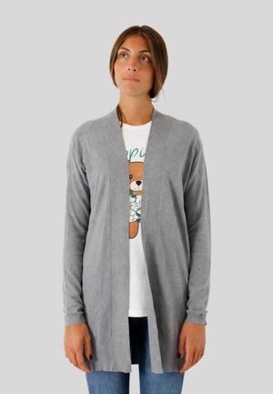 Donna che indossa un cardigan grigio aperto davanti sopra una T-shirt bianca con una stampa di un orsetto e jeans blu, in piedi davanti a uno sfondo semplice.