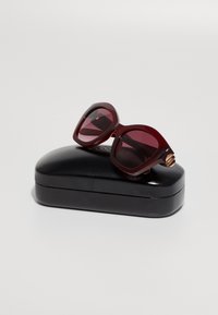 Μπους oversized γυαλιά ηλίου σε μπορντό απόχρωση με γωνιακούς σκελετούς και σκουρόχρωμους φακούς, ακουμπούν σε μια μαύρη σκληρή θήκη. Σημαντικές λεπτομέρειες από ανοξείδωτο ατσάλι.