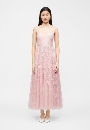 Mlada ženska stoji, oblečena v roza cvetlično čipkasto obleko brez rokavov z oprijetim trupom in tila krilom, kombinirano z belimi koničastimi čevlji.