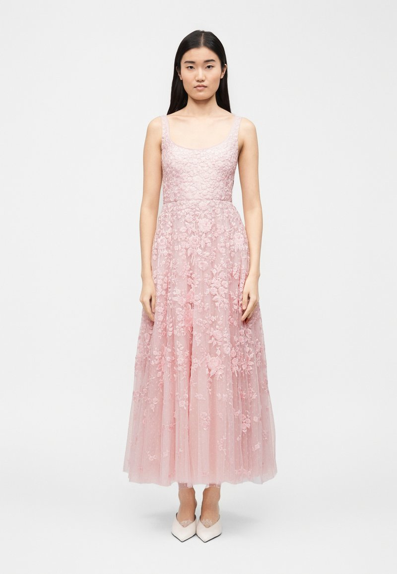 Νεαρή γυναίκα που στέκεται, φοράει ένα αμάνικο ροζ φόρεμα με λουλουδάτη δαντέλα, εφαρμοστή μέση και φούστα από τούλι, συνδυασμένο με λευκά μυτερά παπούτσια.