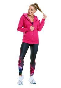 Roze ritsjack met capuchon, gecombineerd met zwarte legging met een kleurrijk geometrisch patroon. Witte sportschoenen maken de outfit compleet.