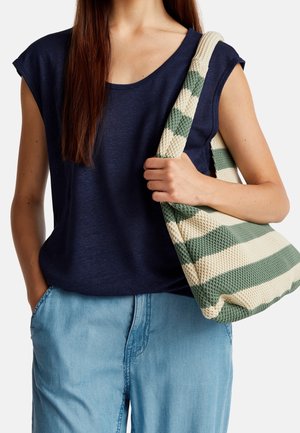 Kobieta ubrana w granatowy, bezrękawnik i jasnoniebieskie dżinsy, trzymająca zielono-kremową paskowaną dzianinową torbę na ramię.