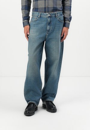 Jean large en denim bleu clair avec une finition délavée, design à cinq poches et fermeture à bouton en métal, associés à des chaussures noires et une chemise à carreaux.
