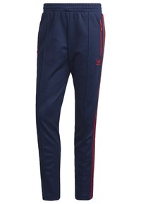 Marinblå atletiska byxor med avslappnad passform, elastisk midja och röda accentstrimmor längs sidorna. Har dragkedjefickor på sidan.