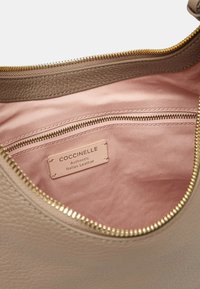 Interno di una borsa a tracolla in pelle beige con rivestimento in tessuto rosa, tasca con zip e etichetta con la dicitura "Coccinelle Authentic Italian Leather." Dettaglio della zip oro.