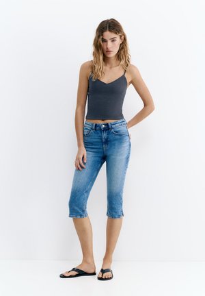 Jeune femme aux cheveux ondulés et lâchés portant un caraco gris foncé, un short denim bleu au genou et des tongs noires, debout devant un fond blanc.