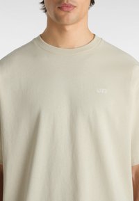 T-shirt a maniche corte color beige chiaro realizzato in cotone morbido. Presenta un piccolo logo ricamato "VANS" in bianco sul lato sinistro del petto. Texture liscia.